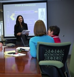 Dr. Shivani Sahni of the Marcus Institute for Aging Research in Boston, MA stands at the head of a conference table, presenting to five colleagues who are seated around the table. Behind Dr. Sahni is a screen where a slide with the title Protein &amp; Osteoporosis is being projected.