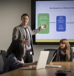 A researcher at the Marcus Institute for Aging Research in Boston, MA stands in front of a screen displaying a presentation. Other researchers are sitting around a table as he speaks and gestures toward them.